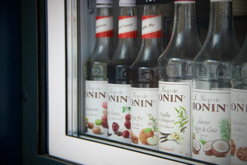 pexels-photo-12482965-12482965 Close-up of Monin syrup bottles displayed in a bar window showcasing flavors like apple pie and coconut.